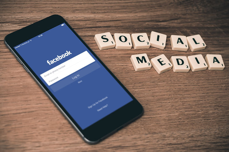A mobile phone on a wooden desk displays the app Facebook. Next to it are scrabble tiles that spell the words Social Media.